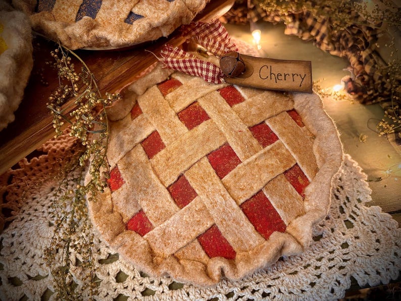 K&ouml;nnte beinhalten: Ein Filz-Kirschkuchen-Ornament mit einem Gitterkrustendesign. Der Kuchen hat eine rote F&uuml;llung und ein Holzschild mit der Aufschrift "Cherry". Eine dekorative Glocke und ein Vichy-Band sind am Schild befestigt. Das Ornament befindet sich auf einer wei&szlig;en Spitzendecke.