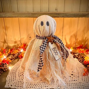 May include: A beige fabric ghost decoration with black eyes and a black and white checkered scarf. A wooden star and a tag that says "BOO" are attached. The ghost is sitting on a white crocheted doily, with fall leaves and lights in the background.