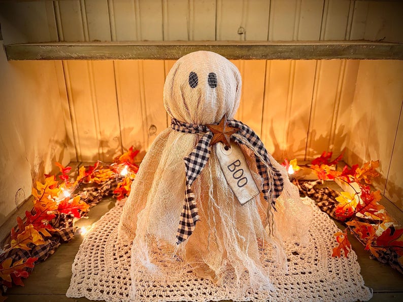 May include: A beige fabric ghost decoration with black eyes and a black and white checkered scarf. A wooden star and a tag that says "BOO" are attached. The ghost is sitting on a white crocheted doily, with fall leaves and lights in the background.