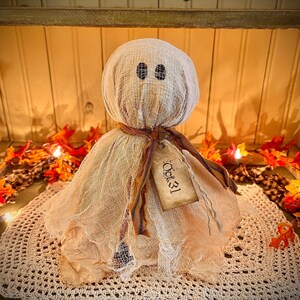May include: A handmade Halloween ghost decoration. The ghost is made of beige fabric with black eye details and a tag that reads "Oct. 31". The ghost is sitting on a white crocheted doily, surrounded by fall leaves.