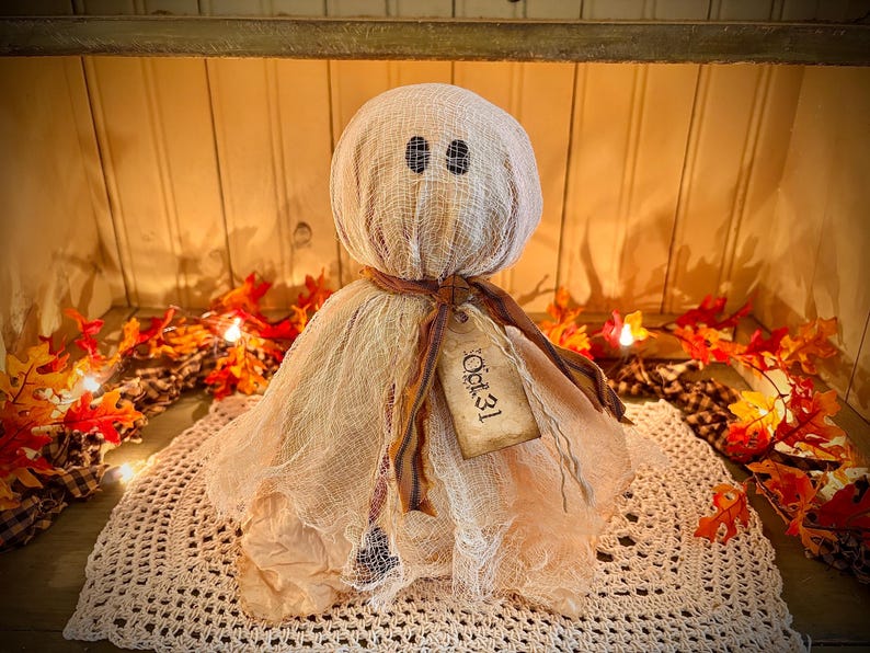 May include: A handmade Halloween ghost decoration. The ghost is made of beige fabric with black eye details and a tag that reads "Oct. 31". The ghost is sitting on a white crocheted doily, surrounded by fall leaves.