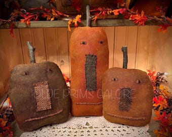 May include: Three fabric pumpkin figures with stitched facial features and burlap patches. The left pumpkin is dark brown, the centre is tall and orange, and the right is light brown. Each has a twig stem and a stitched mouth on the front.