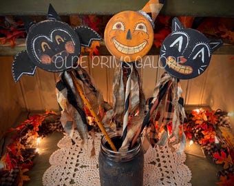 May include: Three decorative wooden sticks featuring painted faces of a black bat, an orange jack-o'-lantern with a plaid hat, and a black cat. Each stick is adorned with strips of frayed fabric in orange, black, and tan, and placed in a dark jar.