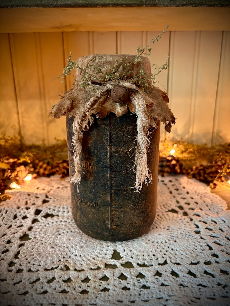 May include: A rustic, brown glass jar with a burlap lid and twine accents. The jar has markings for 250ml and 50ml. The jar is sitting on a white crocheted doily.