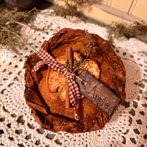 May include: A brown fabric pie crust with a cinnamon stick, dried apple slices, and a red and white checkered ribbon. A brown tag with the word "Apple" is attached to the pie crust. The pie crust is sitting on a white crocheted doily.