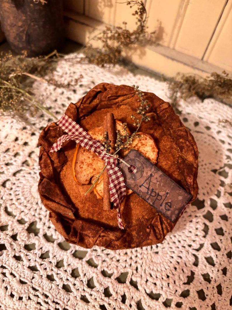 May include: A brown fabric pie crust with a cinnamon stick, dried apple slices, and a red and white checkered ribbon. A brown tag with the word "Apple" is attached to the pie crust. The pie crust is sitting on a white crocheted doily.