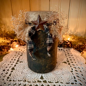 May include: A rustic mason jar with a burlap and plaid bow, a star-shaped metal accent, and dried flowers. The jar is sitting on a white crocheted doily.