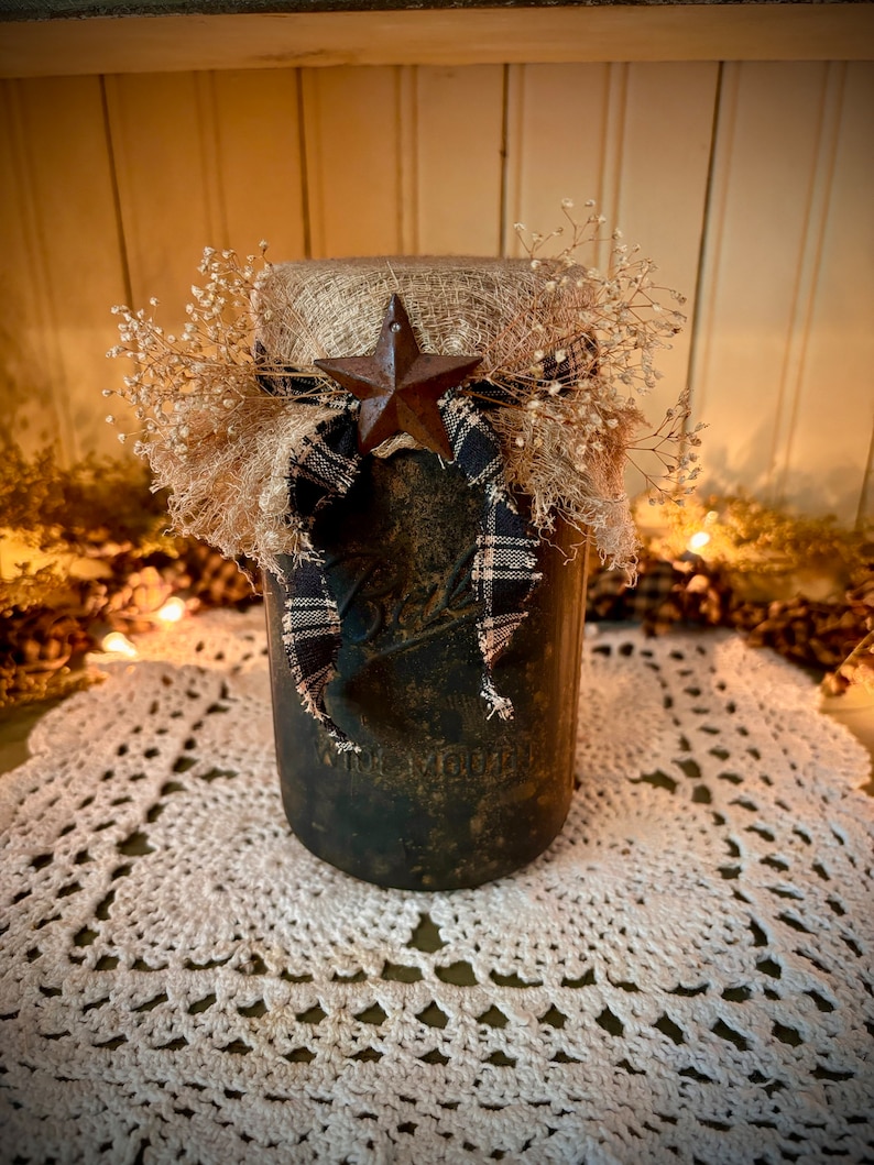 May include: A rustic mason jar with a burlap and plaid bow, a star-shaped metal accent, and dried flowers. The jar is sitting on a white crocheted doily.