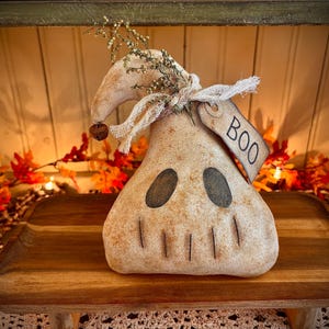 May include: A white ghost-shaped Halloween decoration with a burlap bow and a tag that says "BOO". The ghost is sitting on a wooden shelf with autumn leaves and lights.