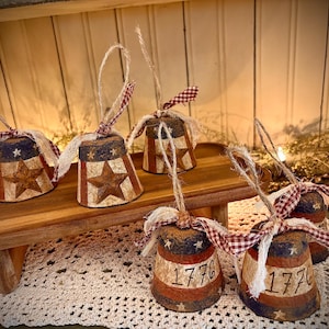 May include: Six small, hand-painted, wooden bells with red, white, and blue patriotic designs. The bells are decorated with stars and the number 1776. The bells are hanging from a wooden shelf.