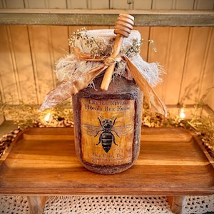Peut inclure: Un pot de miel rustique avec un dipper en bois. Le pot est enveloppé de toile de jute et noué avec un ruban. L'étiquette indique "Little River Honey Bee Farm" avec une illustration d'abeille. Le pot est posé sur un plateau en bois.