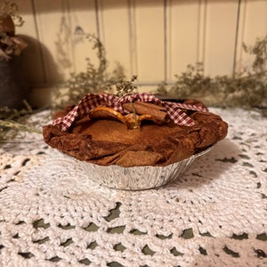May include: A small, brown pie with a red and white checkered fabric top. The pie is in a silver foil pie pan and is sitting on a white crocheted doily.