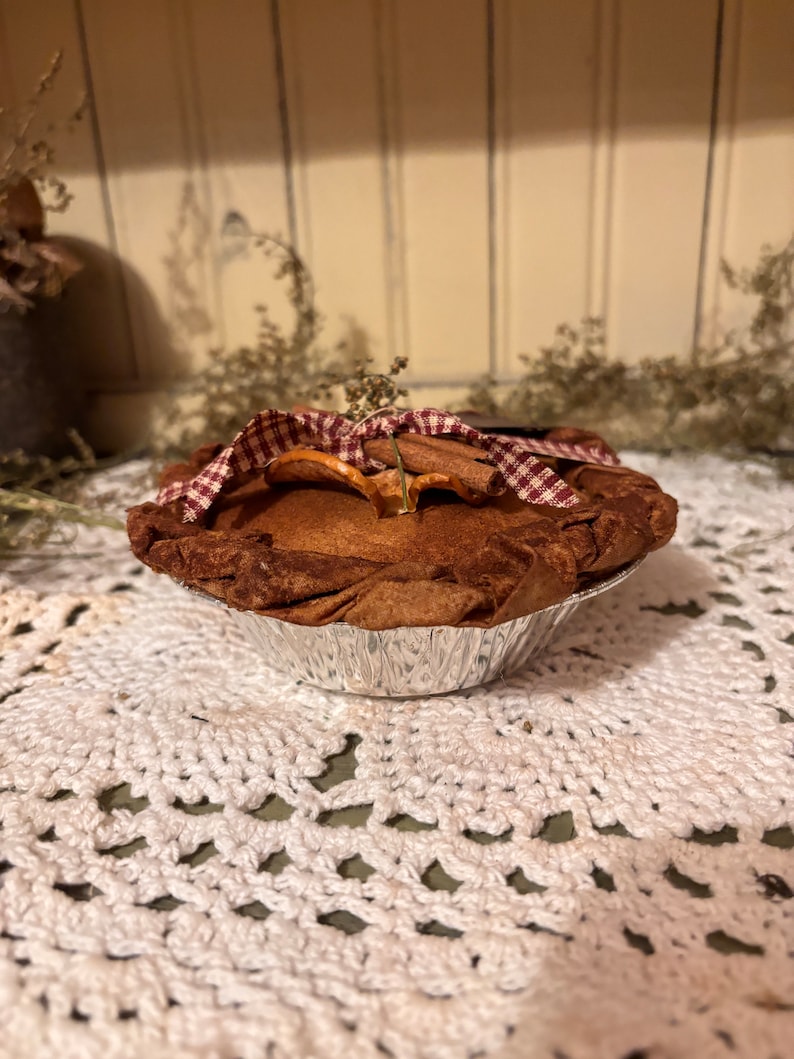 May include: A small, brown pie with a red and white checkered fabric top. The pie is in a silver foil pie pan and is sitting on a white crocheted doily.