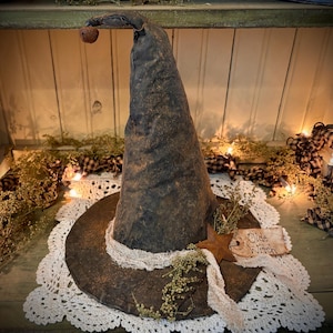 May include: A large, black witch's hat with a brown brim and a bell on top. The hat is sitting on a white crocheted doily with dried flowers and a sign that says "Olde Witch".