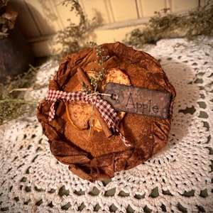 May include: A brown fabric apple pie with a cinnamon stick and a red and white gingham ribbon. The pie is labeled "Apple" and is sitting on a white crocheted doily.