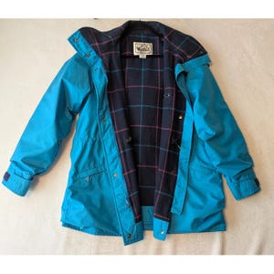 May include: A bright turquoise jacket featuring a dark navy blue interior lining with a red and teal plaid pattern. The jacket includes a front zipper, snap button closures, and a label at the neck that reads Woolrich with a sheep logo.