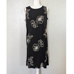 May include: A sleeveless black dress with a floral pattern. The dress features a scoop neckline and a knee-length hemline. The floral design is in a cream color, creating a contrast against the black fabric. The dress is displayed on a mannequin.