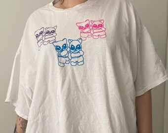 May include: White oversized t-shirt with a graphic of blue and pink cartoon raccoons holding hands.