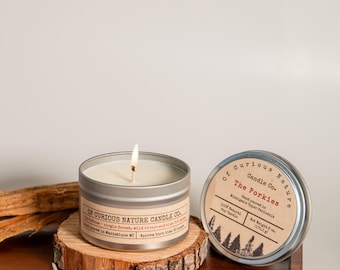 May include: A lit candle in a silver tin with a label that reads "Of Curious Nature Candle Co." sits on a wooden slice. The lid of a second candle is visible, with text that reads "The Porkies".