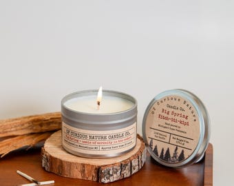 May include: A lit candle in a silver tin, labeled "Big Spring" by Of Curious Nature Candle Co., sits on a wooden slice. A second tin with the same label is next to it. The candle is 6 oz.