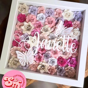 May include: A white frame filled with paper flowers in shades of pink, cream, and purple. The name "Jaurelle" is written in white script across the flowers. Two white paper butterflies are also included in the design.
