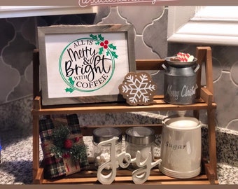 Könnte beinhalten: Ein zweistufiges Holzregal mit einem weißen Schild, auf dem "All is Merry & Bright with Coffee" steht, und einem Schneeflocken-Anhänger. Das Regal enthält außerdem eine Milchkanne aus Metall mit der Aufschrift "Merry Christmas", eine kleine Geschenkbox, zwei Glasbehälter, einen weißen Zuckerbehälter aus Keramik und ein weißes Holzschild mit der Aufschrift "Joy".