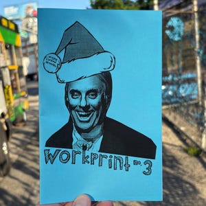 May include: A blue print with a black and white image of a smiling man wearing a Santa hat. The print has the text "MISFIT CINEMA JOURNAL" on the hat and "WORKPRINT no. 3" below the image.