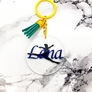 May include: A clear acrylic keychain with a gold chain and a green tassel. The keychain features a silhouette of a gymnast and the name "Lena" in blue lettering.