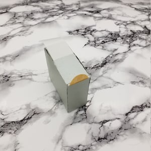 Könnte beinhalten: Eine kleine, hellgraue Schachtel mit einem weißen Deckel und einem goldenen Aufkleber an der Seite.