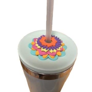 K&ouml;nnte beinhalten: Ein hellgrauer Becher mit einem Blumendesign auf dem Deckel. Der Deckel ist hellgr&uuml;n mit einem bunten Blumendesign in Orange, Lila, Rosa und Blau. Ein grauer Strohhalm ist in den Deckel gesteckt.
