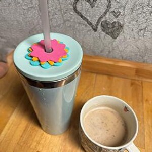 K&ouml;nnte beinhalten: Ein hellblauer isolierter Becher mit einem Blumendeckel und einem Strohhalm, neben einer Tasse mit Blumenmuster, die mit einem Getr&auml;nk gef&uuml;llt ist. Der Becher steht auf einer Holzoberfl&auml;che, mit einer Herzzeichnung im Hintergrund.