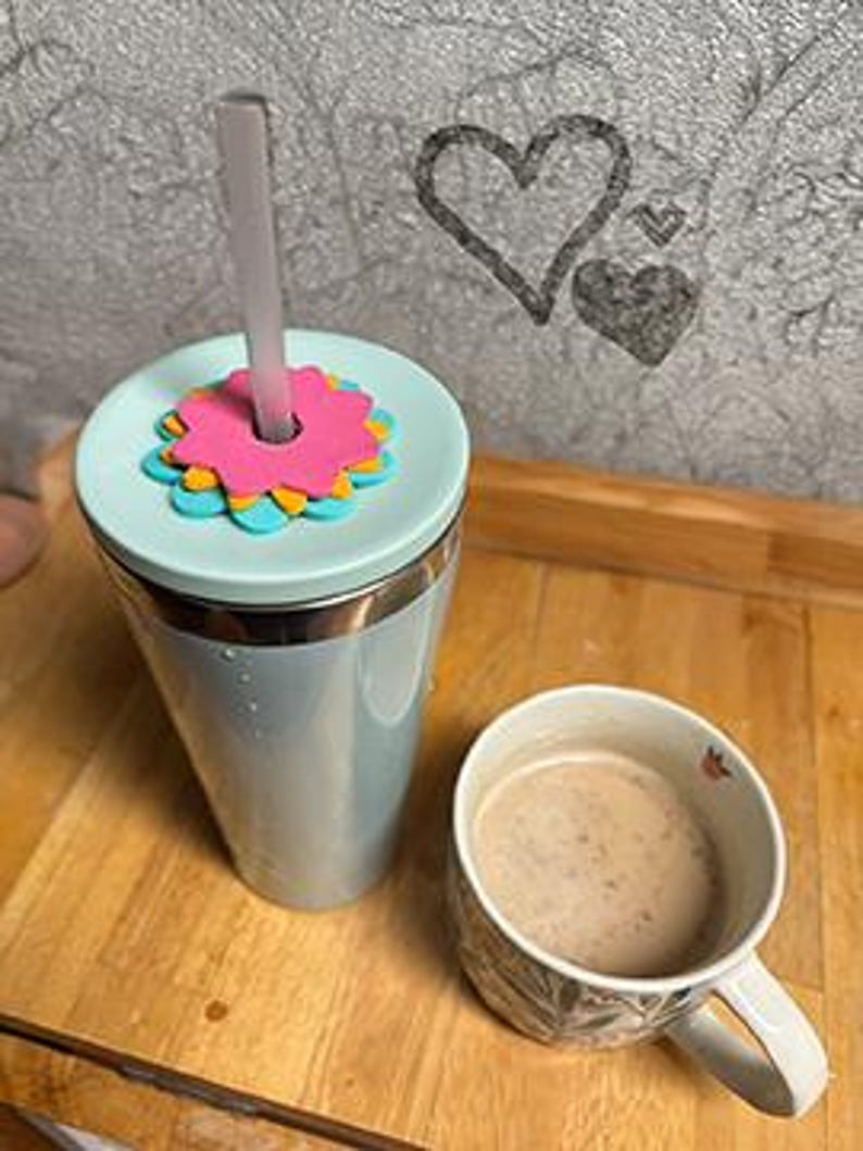 K&ouml;nnte beinhalten: Ein hellblauer isolierter Becher mit einem Blumendeckel und einem Strohhalm, neben einer Tasse mit Blumenmuster, die mit einem Getr&auml;nk gef&uuml;llt ist. Der Becher steht auf einer Holzoberfl&auml;che, mit einer Herzzeichnung im Hintergrund.