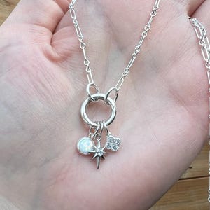 May include: A silver necklace with a large circular pendant and multiple charms. The charms include a white opal-like sphere, a starburst, and a four-leaf clover. The necklace is held in a hand.