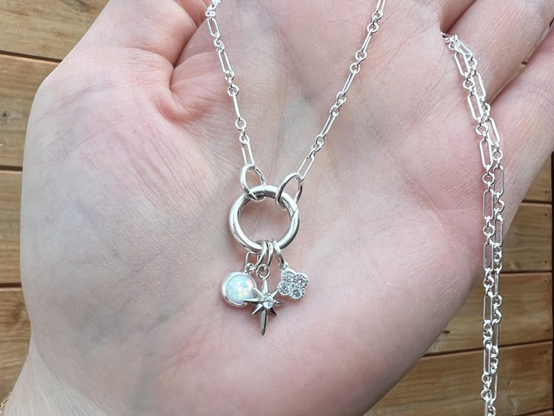 May include: A silver necklace with a large circular pendant and multiple charms. The charms include a white opal-like sphere, a starburst, and a four-leaf clover. The necklace is held in a hand.