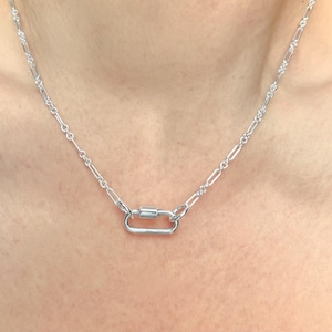 May include: A silver-colored necklace with a carabiner clasp. The necklace chain has a delicate, elongated link design. The clasp is a rectangular shape with a cylindrical bar in the center. The necklace is displayed against a skin-toned background.
