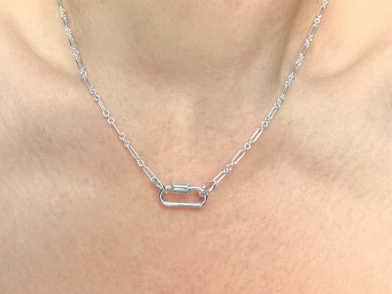 May include: A silver-colored necklace with a carabiner clasp. The necklace chain has a delicate, elongated link design. The clasp is a rectangular shape with a cylindrical bar in the center. The necklace is displayed against a skin-toned background.
