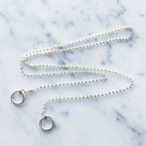May include: A silver chain necklace with two silver rings attached. The chain is on a white marble surface.