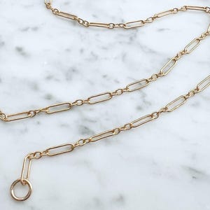 May include: Gold-colored chain necklace with elongated oval links and circular clasps. The jewelry is displayed on a white and gray marble surface. The chain is delicate and appears to be made of metal.