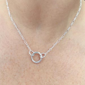 May include: A delicate silver necklace featuring a small, centered circular pendant. The necklace has a delicate chain with elongated links. The jewelry is displayed on a person's neck, showcasing its simple elegance.