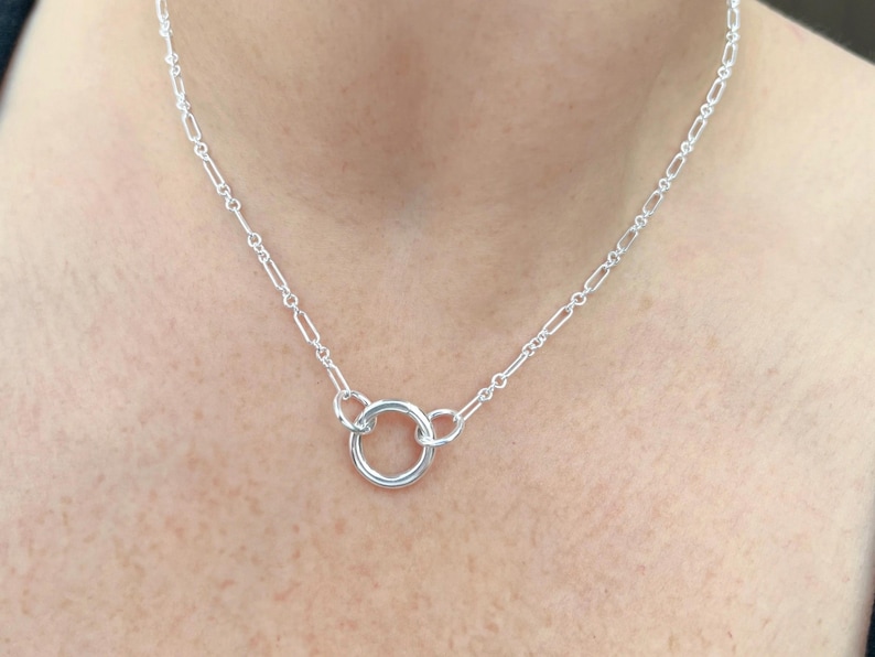 May include: A delicate silver necklace featuring a small, centered circular pendant. The necklace has a delicate chain with elongated links. The jewelry is displayed on a person's neck, showcasing its simple elegance.