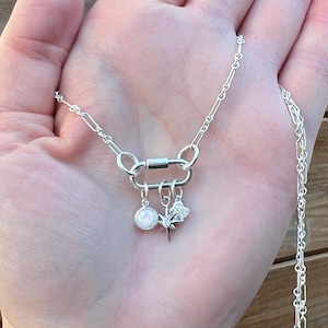 May include: A silver necklace with a unique clasp and three charms. The charms include a round opal-like stone, a starburst, and a floral design. The necklace is displayed on a hand.