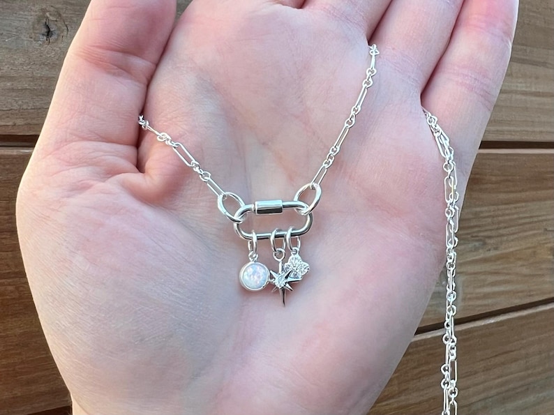 May include: A silver necklace with a unique clasp and three charms. The charms include a round opal-like stone, a starburst, and a floral design. The necklace is displayed on a hand.