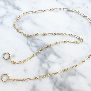 May include: A delicate gold-colored chain necklace with a rectangular link design. The necklace has two circular clasps at the ends. The jewelry is displayed on a white and gray marble surface.