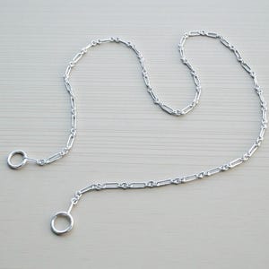 May include: A silver-colored chain with a series of interconnected rectangular links. The chain has a ring at each end. The chain is displayed on a light gray surface. The chain is likely used for jewelry or decorative purposes.