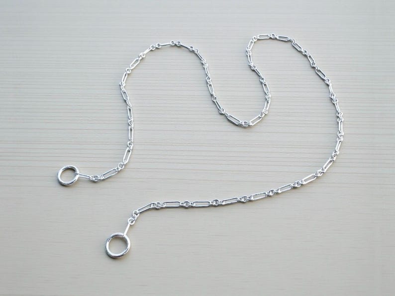 May include: A silver-colored chain with a series of interconnected rectangular links. The chain has a ring at each end. The chain is displayed on a light gray surface. The chain is likely used for jewelry or decorative purposes.