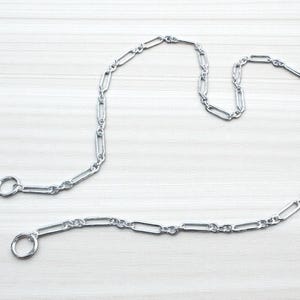 May include: A silver-colored chain with elongated oval links and two circular rings at the ends. The chain is laid out on a light-colored wooden surface. The chain is likely used for jewelry or as a decorative element.