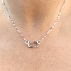 May include: A silver necklace featuring a delicate chain with a rectangular pendant. The necklace is made of a shiny, metallic material and is displayed against a skin tone background. The chain is composed of small, elongated links.