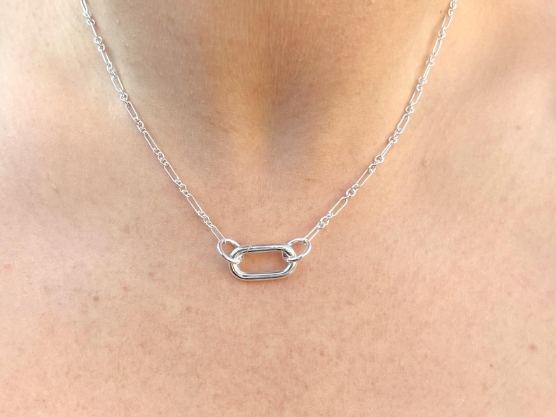 May include: A silver necklace featuring a delicate chain with a rectangular pendant. The necklace is made of a shiny, metallic material and is displayed against a skin tone background. The chain is composed of small, elongated links.