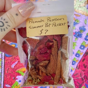 May include: A clear plastic bag filled with dried rose petals, cinnamon sticks, and other herbs. A small yellow tag reads "Premade Passion Simmer Pot Packet". The bag is held against a colorful patterned background.