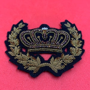 May include: A gold and brown embroidered crown with a wreath of leaves around it. The crown is made of beads and the leaves are made of thread. The patch is on a red background.